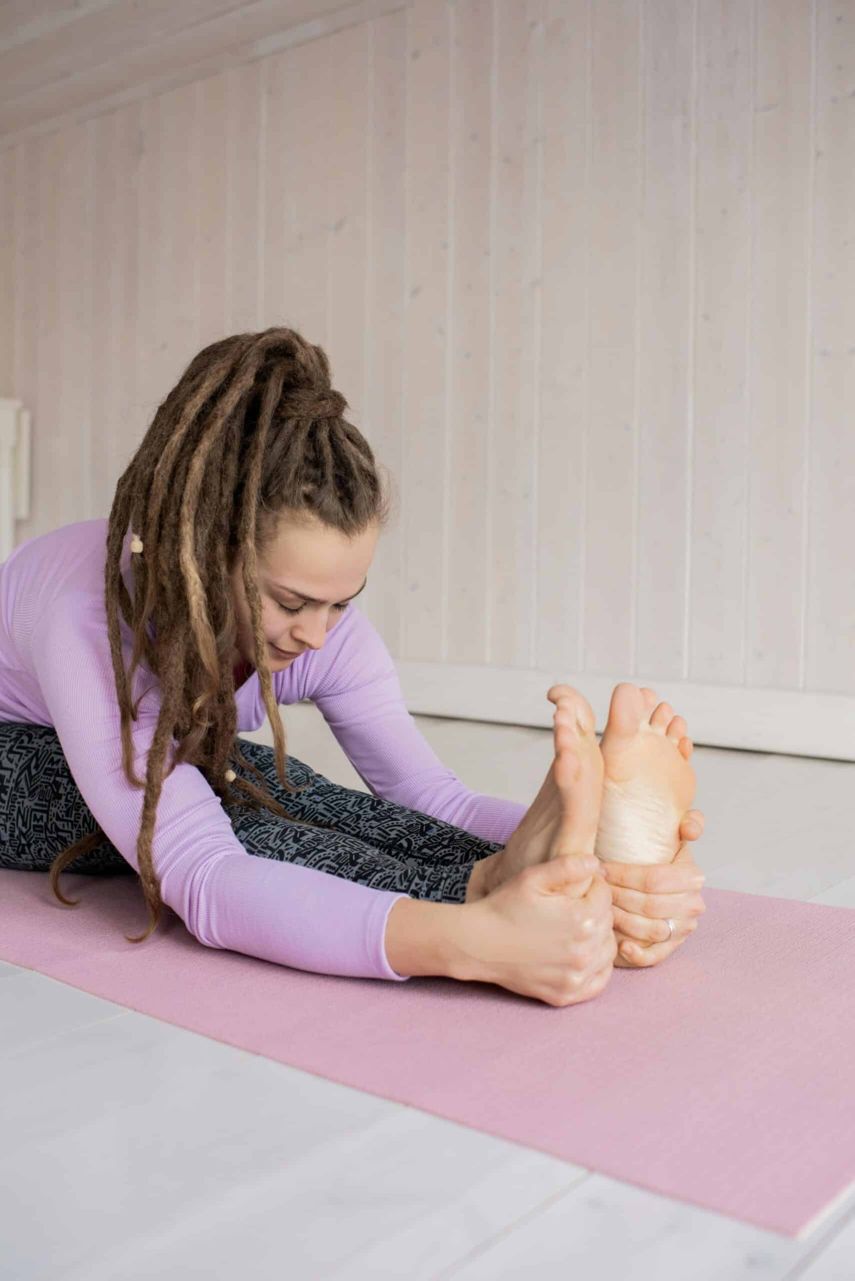 Kobieta z dredami w pozycji skłonu do przodu, trzymająca stopy na macie