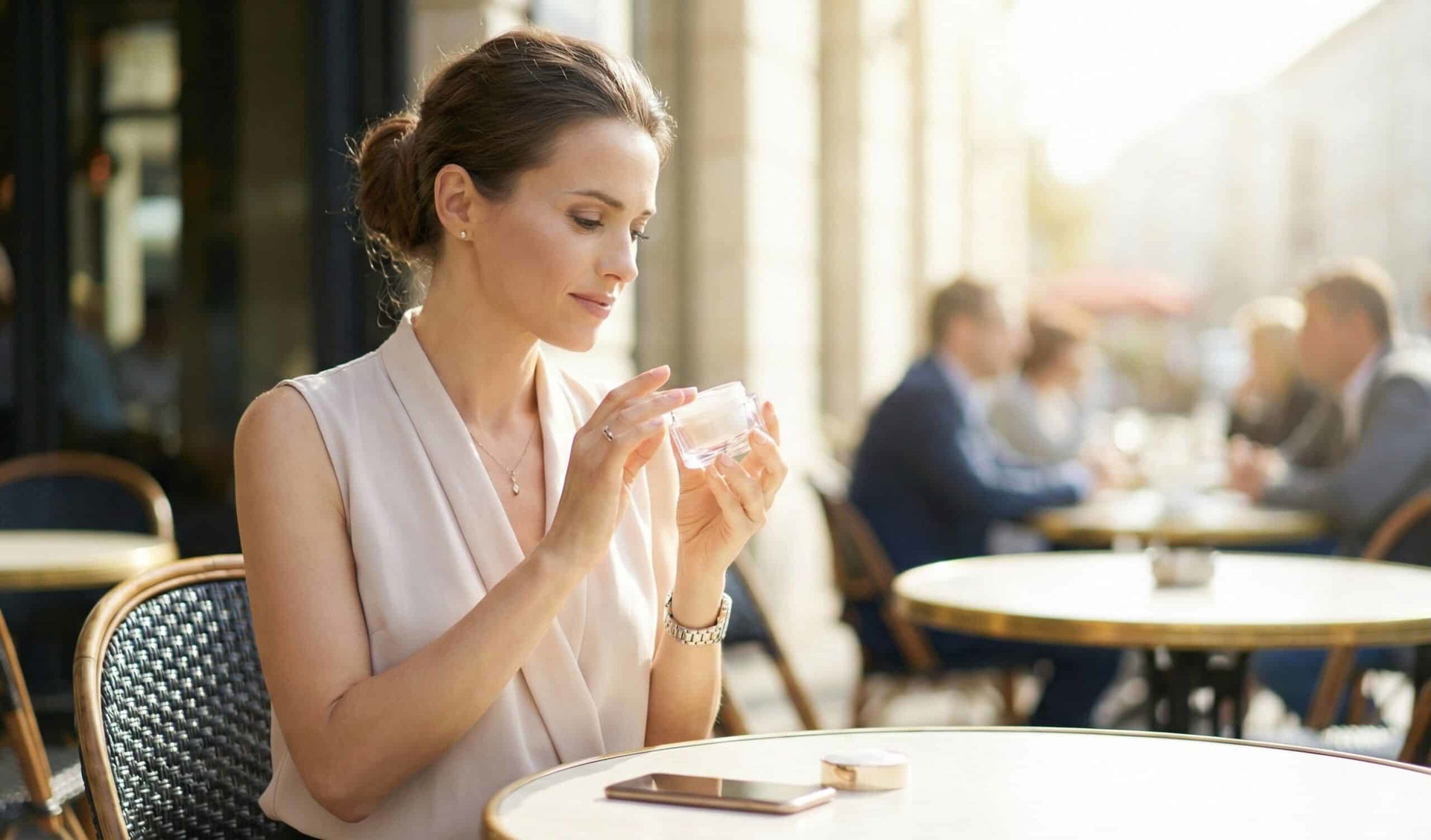 Elegancka kobieta siedząca przy stoliku w kawiarni na świeżym powietrzu, trzymająca w dłoniach otwarty słoiczek kremu, przyglądająca się mu z zainteresowaniem.